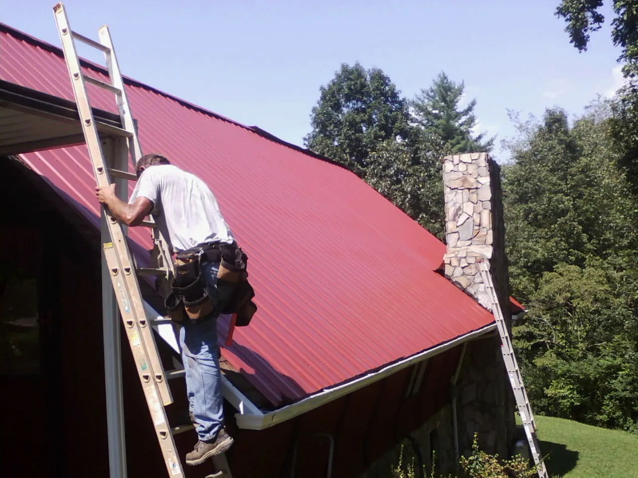 Roofer on a ladder beside a red metal roof and stone chimney — Soffit & Fascia Repair by Roofing Brothers in Grantville